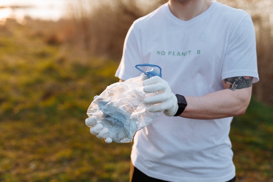 A person wearing a white t-shirt with the text 'NO PLANT B' and a black wristwatch is standing outdoors on a grassy area with blurred brown and green foliage in the background. They are holding a tran