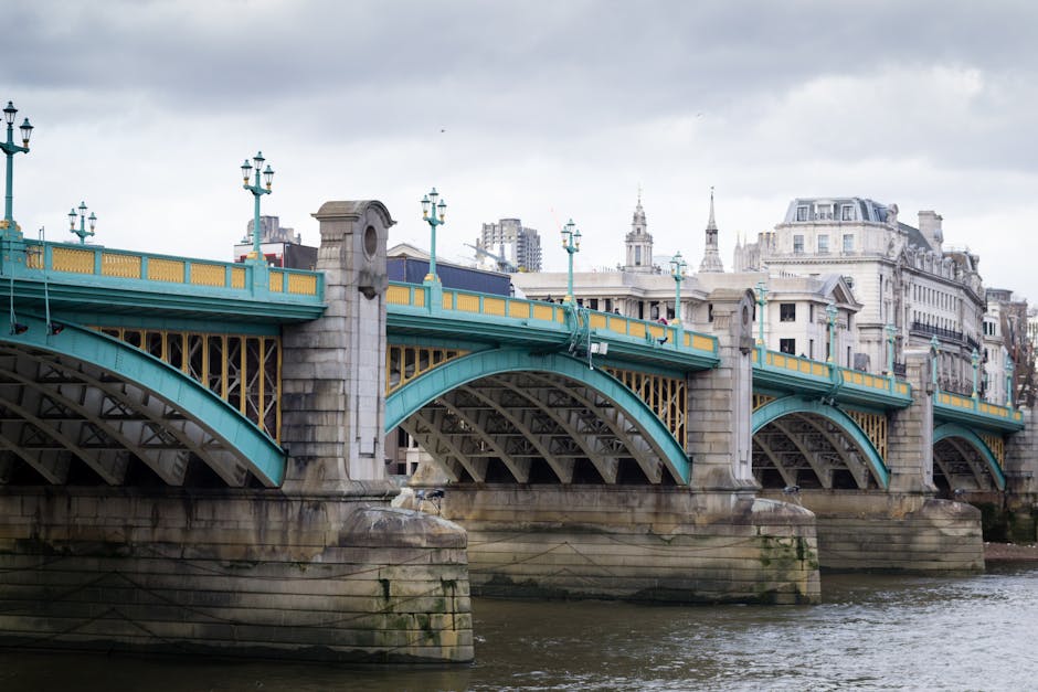 A large white suspension bridge spans across a river under a cloudy sky, with two tall towers connected by numerous cables. The bridge features ornate decorative details and is supported by substantia